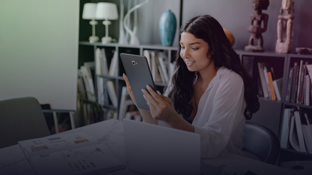 Frau arbeitet mit Tablet, Laptop und Unterlagen am Schreibtisch.