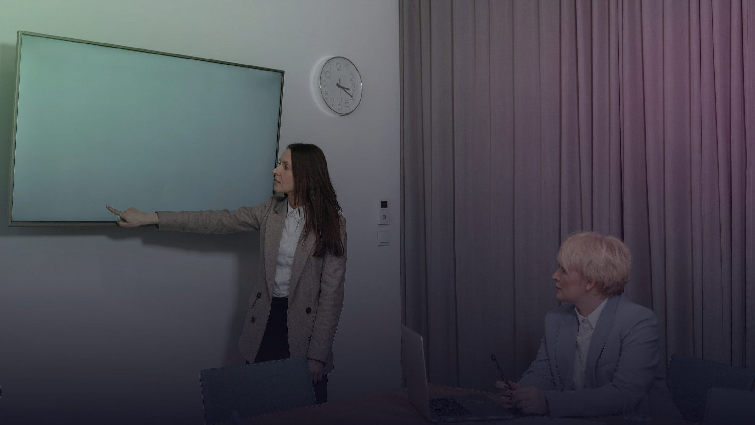 A woman points to a digital board while another woman sits at a table.